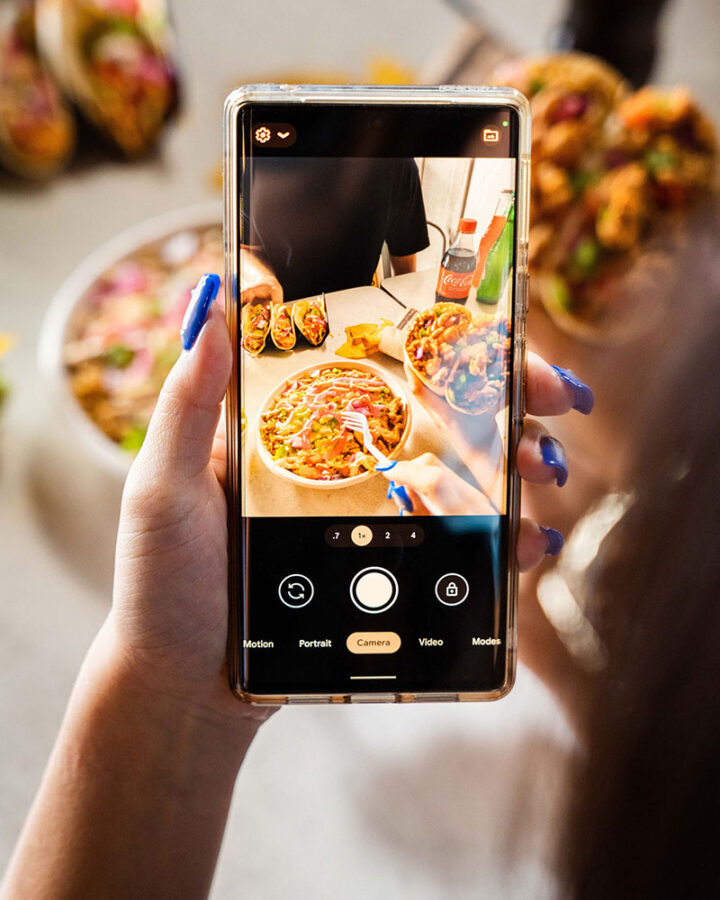 photo of a woman with bright blue nail polish holding her phone and taking a photo of her meal